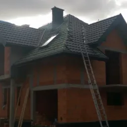 Dom w trakcie budowy, z częściowo położonym, ciemnym dachem i oknem dachowym, widoczna drabina oparta o ścianę, stan surowy otwarty.