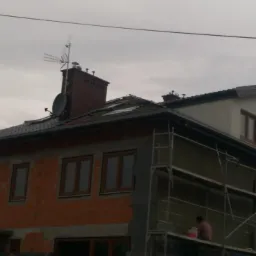 Dwupiętrowy dom w trakcie prac remontowych elewacji i dachu, z robotnikami na rusztowaniu i na dachu, widoczny komin z anteną satelitarną i telewizyjną.