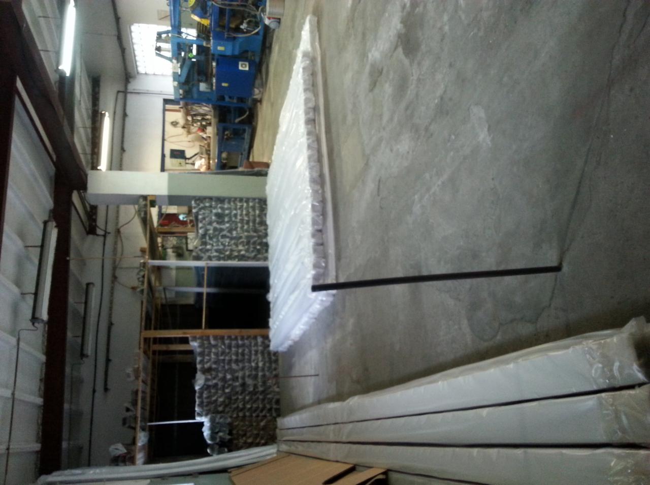 Stosy wełny mineralnej w przezroczystych opakowaniach, ułożone w magazynie z betonową podłogą. W tle maszyna produkcyjna i regały z materiałami.