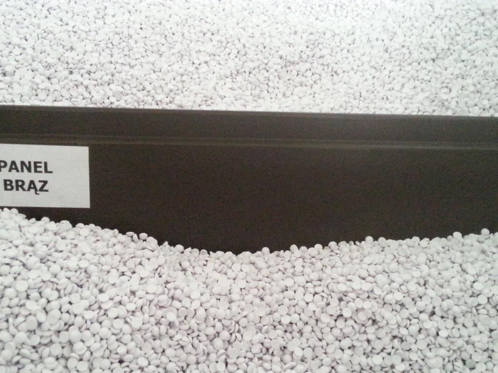 Brązowy panel na tle białych granulek styropianu, z etykietą 'Panel Brąz'. Widok z bliska, prezentujący strukturę materiałów.