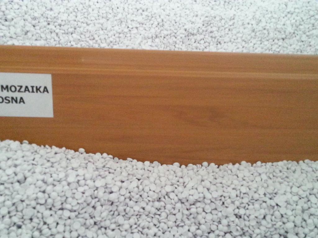Brązowa listwa przypodłogowa z napisem 'Mozaika Osna' otoczona białymi granulkami, przypominającymi styropian. Widoczne nierównomierne oświetlenie.