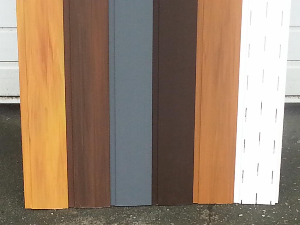 Paleta kolorów paneli elewacyjnych z różnymi fakturami, od jasnego drewna po szary i biały z perforacją, na tle szarej powierzchni.