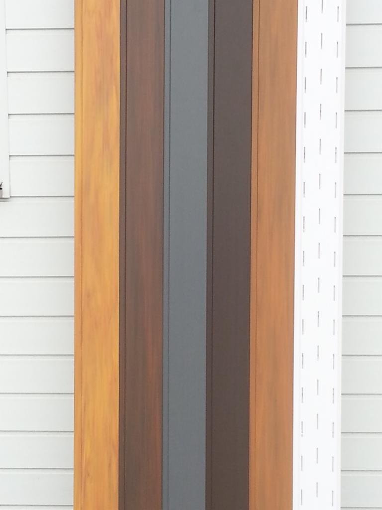 Próbki elewacyjne w odcieniach brązu, szarości i bieli, prezentowane na tle białej ściany z poziomym wzorem. Widoczne różne faktury i kolory materiałów elewacyjnych.