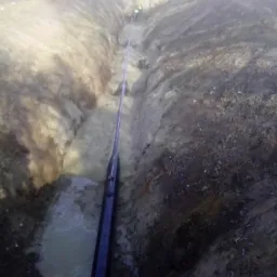 Wykop z ułożonym rurociągiem w ziemi, widoczni pracownicy w oddali. W wykopie woda. Grunt piaszczysto-gliniasty. Prace ziemne w trakcie realizacji.