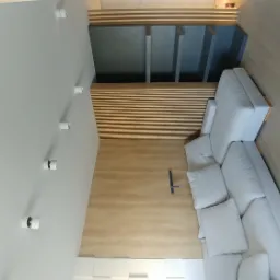 Nowoczesny salon z drewnianą podłogą, panelami ściennymi i narożną sofą w jasnych barwach, oświetlony punktowymi lampami.