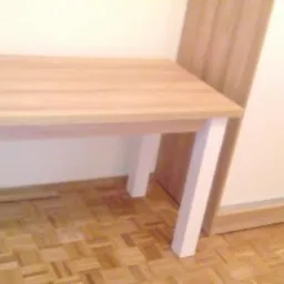 Blat drewniany z białymi nogami na tle podłogi parkietowej i szafy przesuwnej.