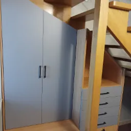 Szara szafa z drewnianą zabudową pod schodami, widoczne szuflady z czarnymi uchwytami i drewniane schody.