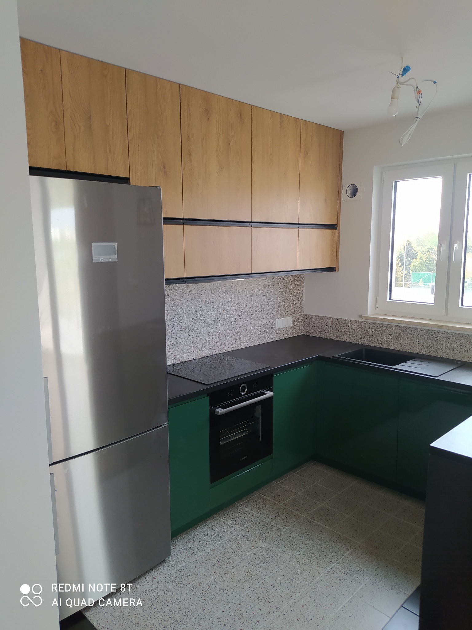 Nowoczesna kuchnia: drewniane szafki górne, zielone szafki dolne, szary blat, srebrna lodówka, płytki mozaikowe na ścianie, niedokończone oświetlenie sufitowe.