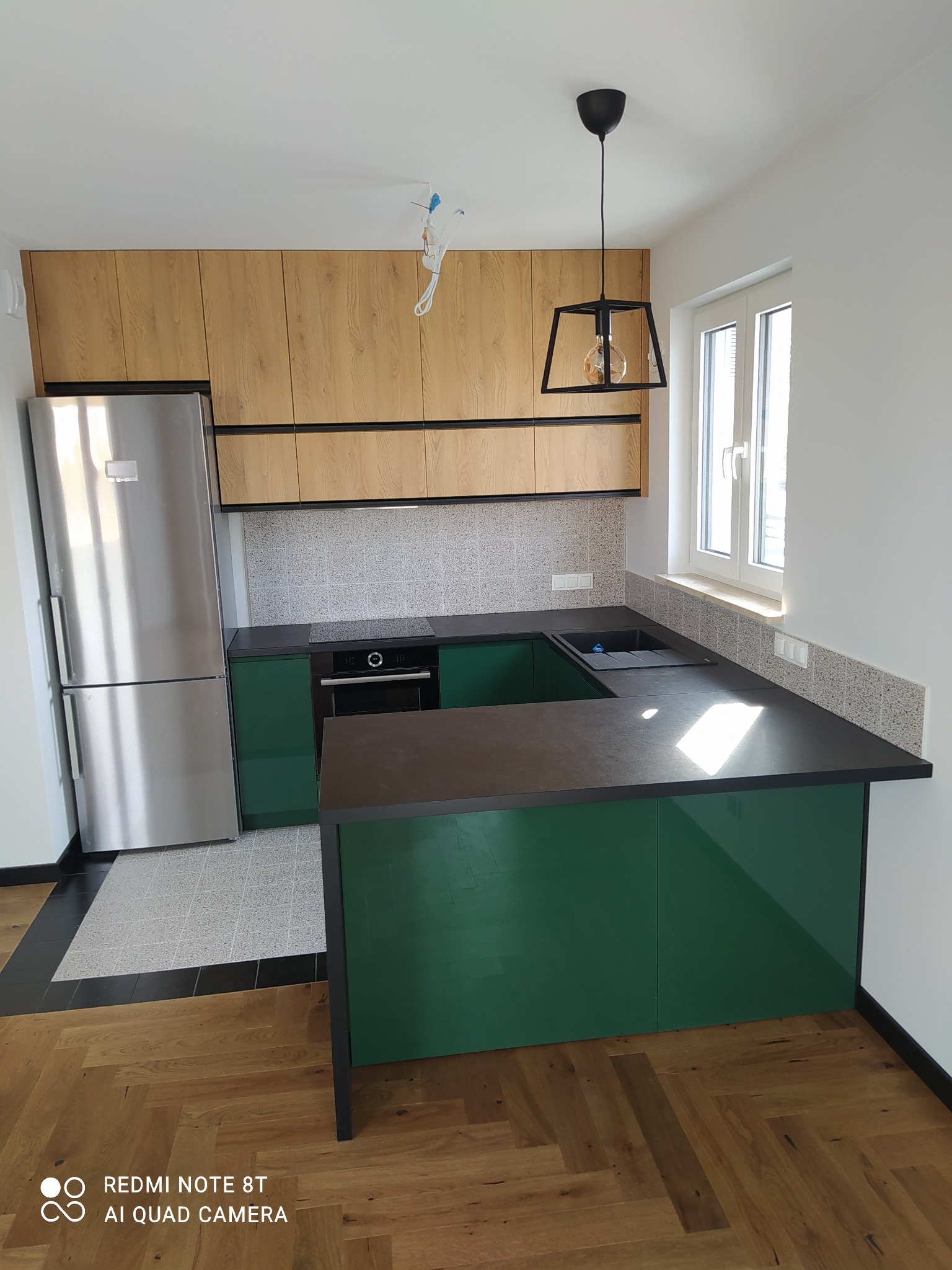 Nowoczesna kuchnia z zielonymi szafkami dolnymi i drewnopodobnymi górnymi, czarnym blatem i podłogą w jodełkę, oświetlona wiszącą lampą geometryczną.