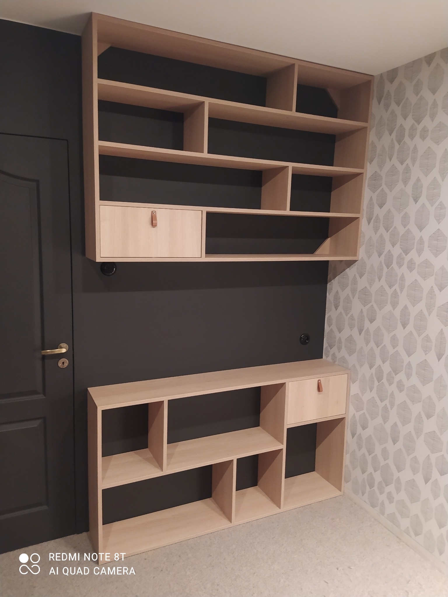 Jasne drewniane regały modułowe z szufladami, zamontowane na ciemnej ścianie z drzwiami i ścianie z wzorzystą tapetą.