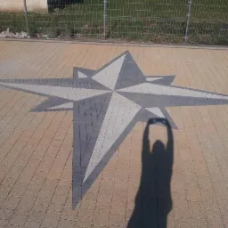 Ułożona kostka brukowa w kształcie gwiazdy kompasu, z cieniem osoby robiącej zdjęcie. Brązowo-szare kostki, siatka ogrodzeniowa w tle.
