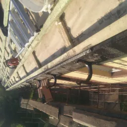 Montaż rynien dachowych z bliska, widoczne haki, deska okapowa zabezpieczona czarną powłoką, drabina oparta o dach. Ujęcie z dołu, lekko obrócone.