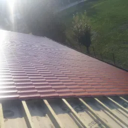 Montaż bordowej blachodachówki modułowej na drewnianej konstrukcji dachu. Widok z góry, w tle zieleń drzew i trawy. Ostre światło słoneczne.