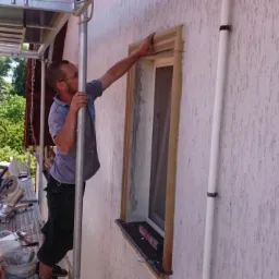 Pracownik na rusztowaniu montuje ozdobne obramowanie okienne na elewacji z tynkiem strukturalnym, widoczne narzędzia i materiały budowlane.