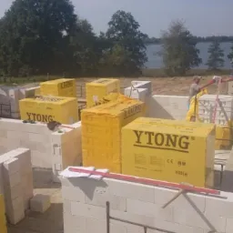 Wznoszenie ścian parterowego domu z bloczków Ytong, widoczne palety z materiałem, pracownicy murują, w tle krajobraz z jeziorem i lasem.