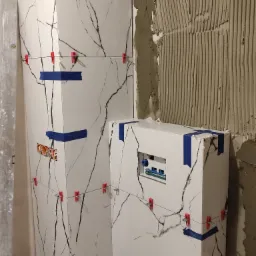 Montaż płytek imitujących marmur na ścianie i zabudowie geberitu, z użyciem niebieskiej taśmy malarskiej i czerwonych klipsów poziomujących, widoczna warstwa kleju na sąsiedniej ścianie.