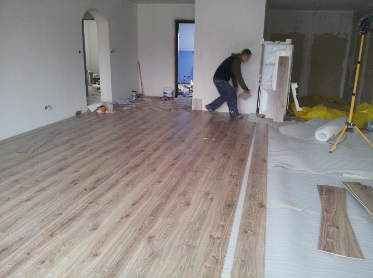 Montaż paneli podłogowych w jasnym odcieniu drewna. Widoczny pracownik układający panele oraz przygotowane materiały i podkład. Częściowo wykończone pomieszczenie.