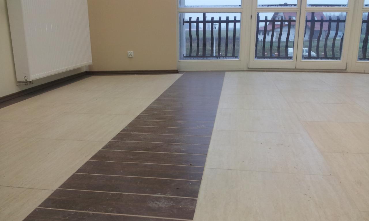 Ułożona podłoga z płytek w odcieniach beżu i brązu, z dekoracyjnym pasem. Widok na okno z czarną balustradą i fragment domu w tle. Grzejnik na ścianie.