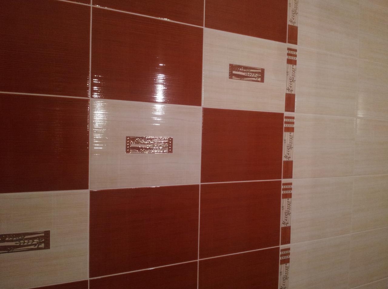 Ściana wyłożona brązowo-beżowymi płytkami z dekoracyjnymi wstawkami. Widoczne fugi i błyszcząca powierzchnia płytek. Całość tworzy eleganckie wykończenie ściany.