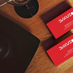 Dwie czerwone wizytówki z białym logo i danymi kontaktowymi, leżące na drewnianym biurku obok czarnego laptopa i kieliszka do wina.