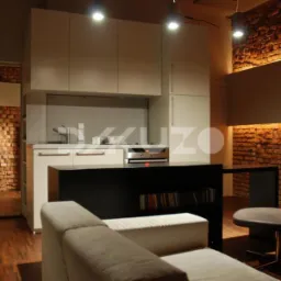 Nowoczesna kuchnia z ceglaną ścianą, oświetleniem punktowym, białymi szafkami i czarną wyspą z regałem na książki, widok z salonu z beżową sofą i krzesłem na metalowej nodze.