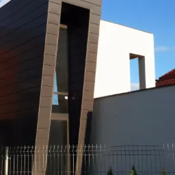 Nowoczesny budynek z ciemną, skośną elewacją i wysokim oknem, widoczny za metalowym ogrodzeniem z młodymi tujami, z fragmentem białej ściany budynku w tle.