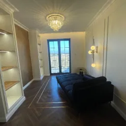 Elegancki pokój z ciemną sofą na parkiecie w jodełkę, oświetlony kryształowym żyrandolem i lampą ścienną z kulistymi kloszami, z zabudowanymi regałami z podświetleniem LED.