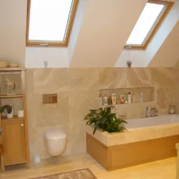 Jasna łazienka na poddaszu z beżowymi płytkami imitującymi marmur, oknami dachowymi i drewnianymi meblami. Uwagę przykuwa wnęka z kosmetykami nad wanną.