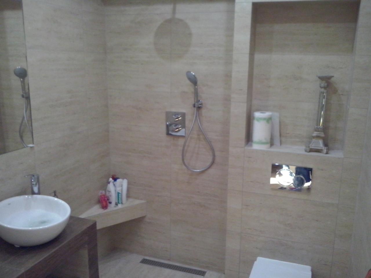 Wykończona łazienka z beżowymi płytkami imitującymi trawertyn, kabiną prysznicową typu walk-in, umywalką nablatową i wnęką z dekoracjami.