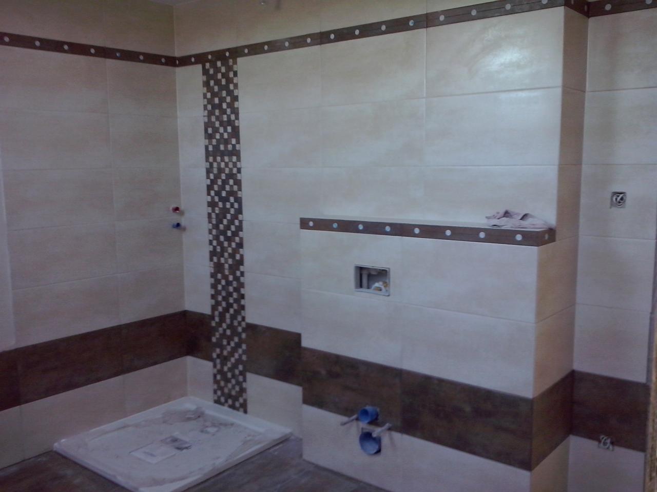 Wykończona łazienka z beżowymi płytkami, brązowymi akcentami i dekoracyjną mozaiką. Widoczne elementy armatury i półka z ręcznikiem. Neutralna kolorystyka.