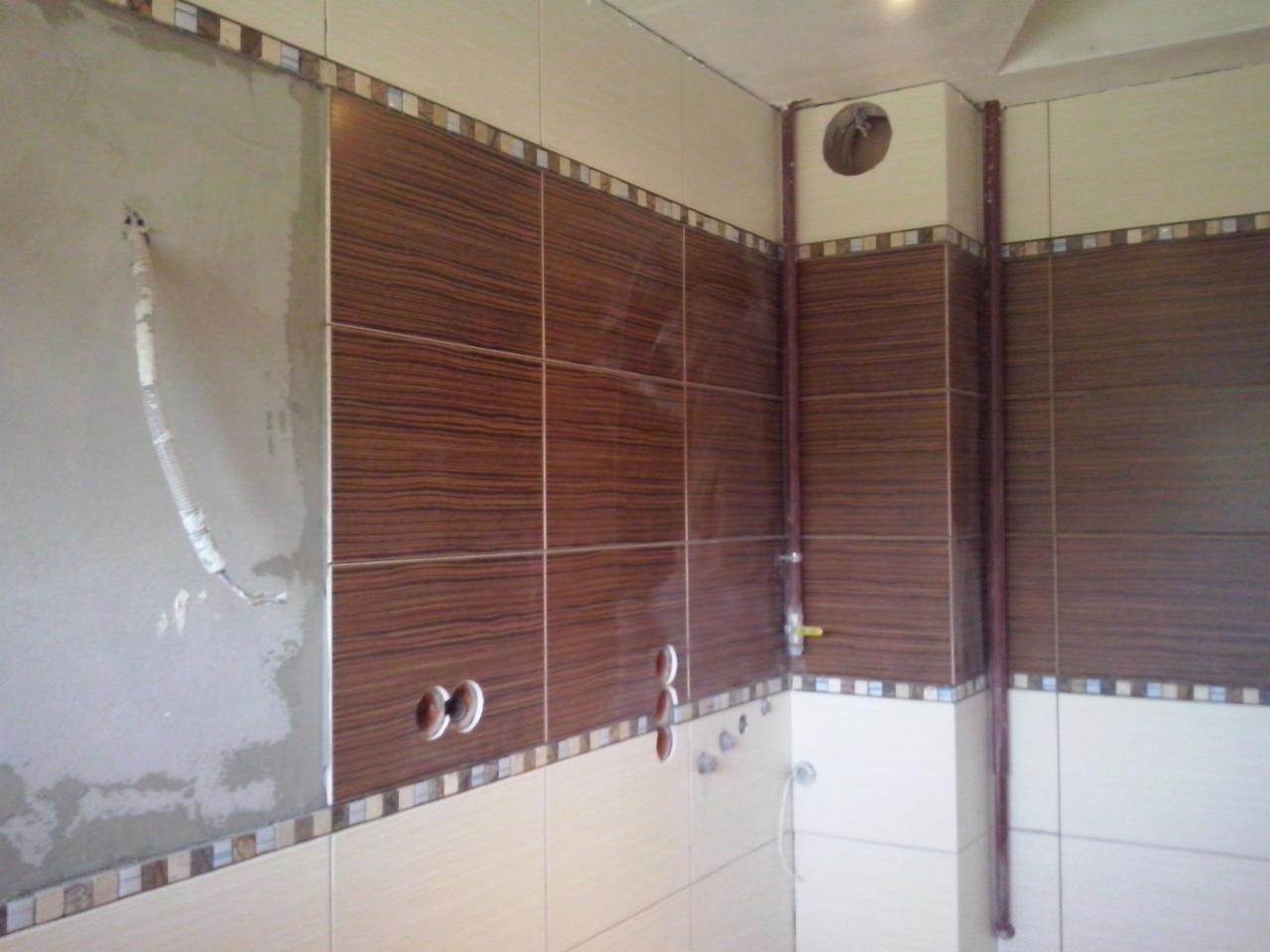 Układanie płytek ściennych w łazience, z widoczną instalacją hydrauliczną i fragmentem nieotynkowanej ściany. Brązowo-beżowa kolorystyka z dekoracyjnym pasem płytek.