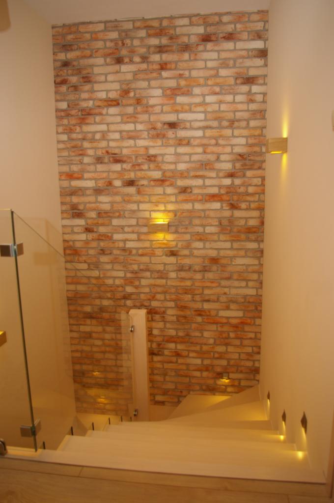 Ściana z cegły dekoracyjnej z oświetleniem punktowym przy schodach z szklaną balustradą. Wykończenie wnętrza w ciepłych barwach.