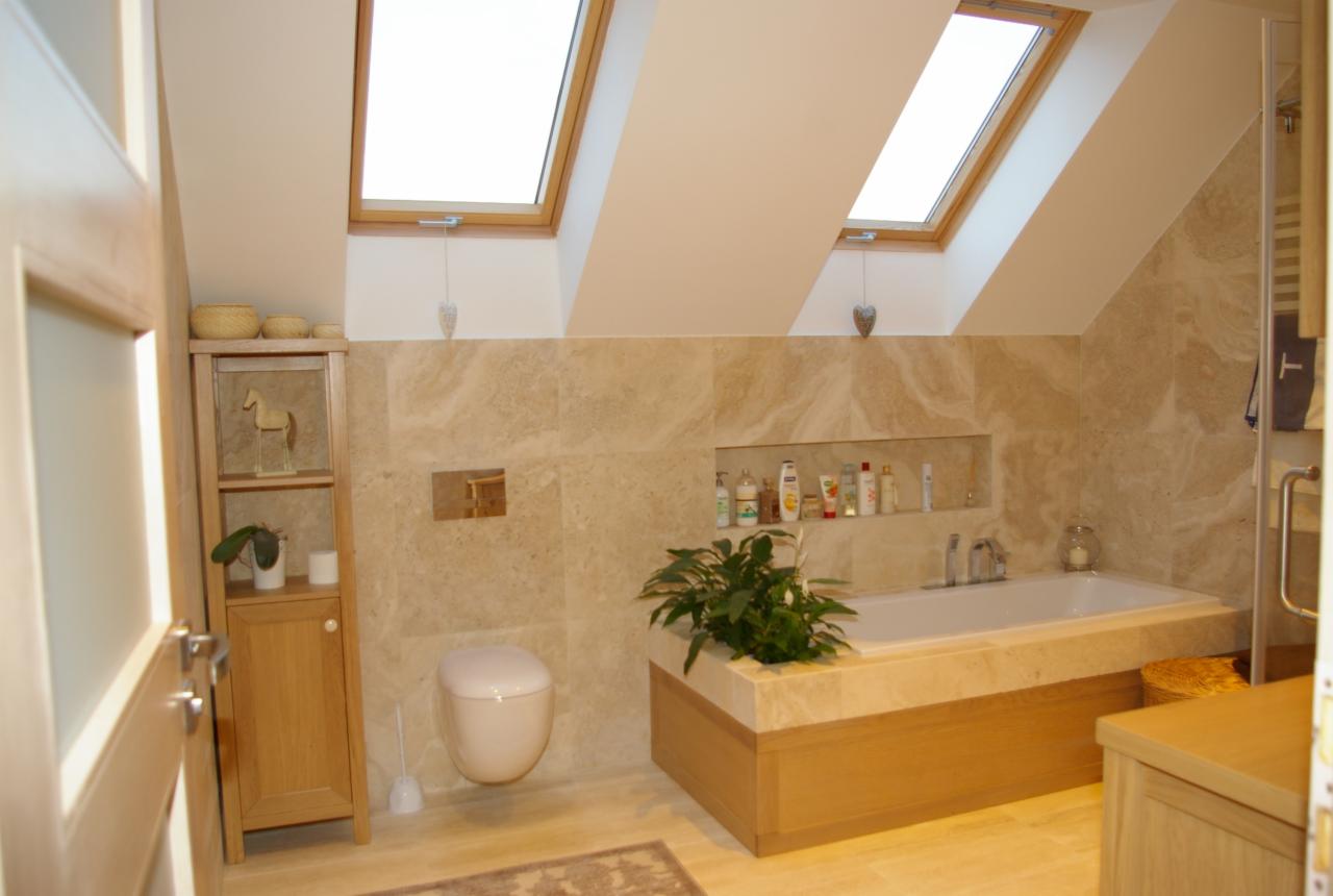 Jasna łazienka na poddaszu z beżowymi płytkami imitującymi marmur, oknami dachowymi i drewnianymi meblami. Uwagę przykuwa wnęka z kosmetykami nad wanną.