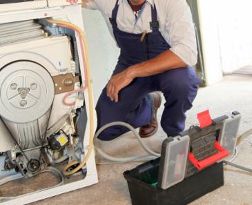 Mężczyzna w kombinezonie naprawia pralkę, z otwartą obudową i widocznymi elementami wewnętrznymi. Obok stoi skrzynka z narzędziami.