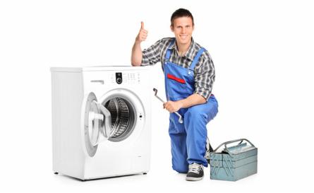 Uśmiechnięty serwisant w kombinezonie z kluczem, kuca obok pralki z otwartymi drzwiami, pokazując kciuk w górę. Obok leży skrzynka narzędziowa.