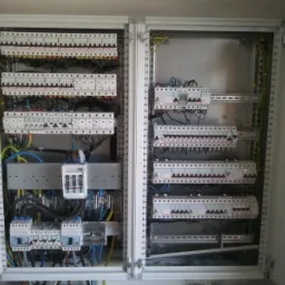 Rozdzielnica elektryczna z licznymi zabezpieczeniami i przewodami wewnątrz szafy, widok z bliska na instalację w metalowej obudowie.