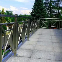 Balkon wykończony szarymi płytkami z nowoczesną, geometryczną konstrukcją ze stali nierdzewnej na tle zieleni i błękitnego nieba.