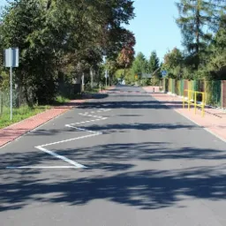 Nowo wyremontowana asfaltowa droga z kostką brukową po bokach, oznakowaniem poziomym w kształcie zygzaka, znakami drogowymi i progami zwalniającymi, otoczona zielenią i zabudową wiejską w słoneczny...