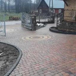 Ułożona nawierzchnia z kostki brukowej w odcieniach czerwieni i szarości, z dekoracyjnymi okręgami z żółtej kostki, prowadząca od ażurowej bramy do schodów wejściowych domu z cegły. Brzeg...