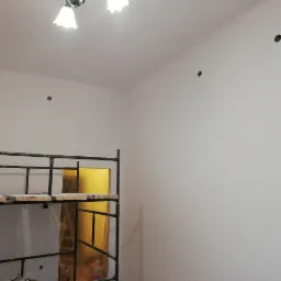 Świeżo pomalowana ściana z widocznymi otworami na gniazdka elektryczne, fragment rusztowania i lampa sufitowa w pomieszczeniu w trakcie remontu.