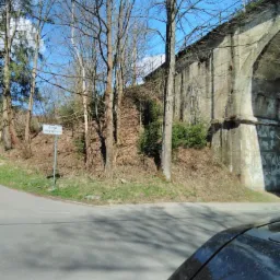 Widok na skrzyżowanie drogi z betonowym wiaduktem kolejowym, z drzewami bez liści i znakiem drogowym. Fragment samochodu w prawym dolnym rogu.