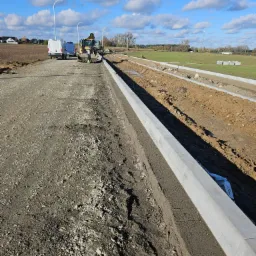 Formowanie krawężnika drogowego z betonu przy użyciu koparki na gruncie, w tle zaparkowane busy i pola uprawne pod błękitnym niebem z chmurami.