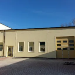 Hala z płyt warstwowych w beżowej kolorystyce, brama garażowa z oknami, widoczne rynny i schody. Na pierwszym planie kostka brukowa.