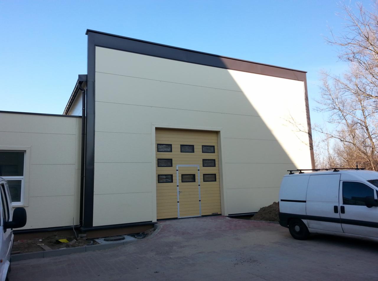 Nowoczesna hala z płyt warstwowych w jasnych barwach, z segmentową bramą garażową i czarnymi obróbkami blacharskimi. Widoczny fragment samochodu dostawczego.
