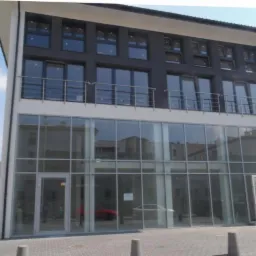 Dwukondygnacyjny budynek biurowy z dużą przeszkloną fasadą na parterze i rzędem okien na piętrze, balkon z metalową balustradą, widok z poziomu chodnika w słoneczny dzień.