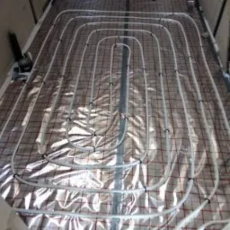 Białe rurki ogrzewania podłogowego ułożone w pętle na srebrnej folii z czerwoną siatką, widoczne szare ściany z betonu komórkowego.