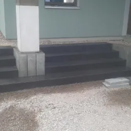 Schody wejściowe z ciemnej kostki brukowej, wykończone jasnymi elementami betonowymi. W tle widoczny budynek i teren przygotowany pod dalsze prace brukarskie.