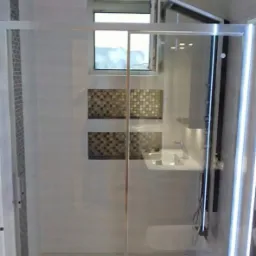 Nowoczesna kabina prysznicowa ze szklanymi drzwiami, mozaiką na ścianie i oknem. Minimalistyczna umywalka z oświetleniem LED. Wykończenie w jasnych, neutralnych kolorach.