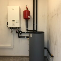 ATOM SYSTEM Tomasz Chrzanowski - Biała jednostka centralna z panelem sterowania Panasonic, czerwony zbiornik wyrównawczy i szary zasobnik ciepłej wody połączone czarnymi rurami w pomieszczeniu z beżowymi ścianami i szarą podłogą.