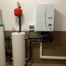 ATOM SYSTEM Tomasz Chrzanowski - Biały zbiornik buforowy z czerwonym naczyniem przeponowym, podłączony do pompy ciepła Ecodan, widoczne rury z izolacją termiczną i osprzęt, na tle brązowych płytek.